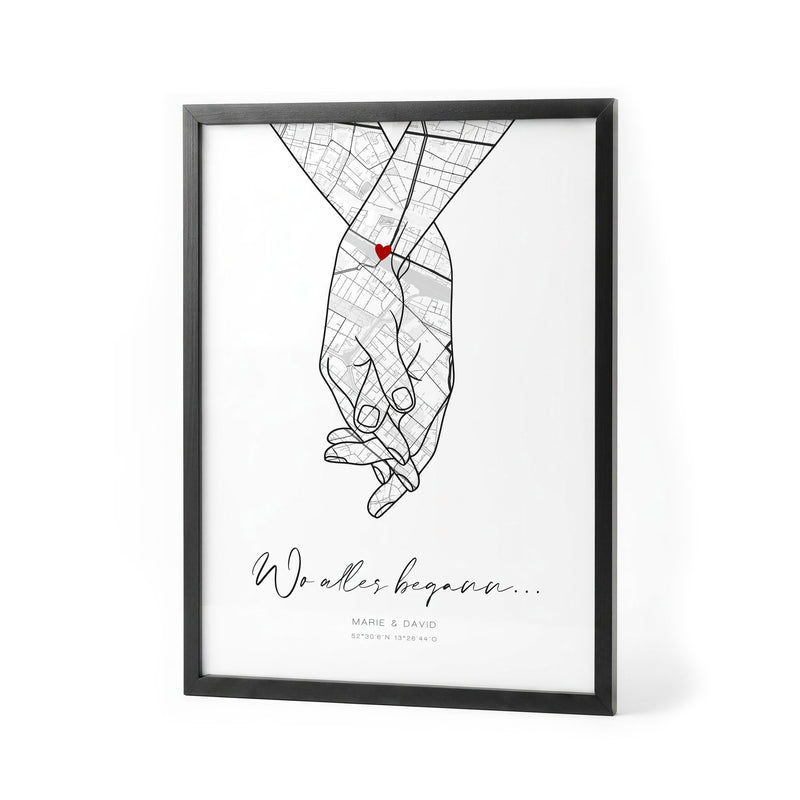 Hand-in-Hand-Kartenposter: Stadtkarte in zwei gezeichneten Händen mit rotem Herz, Titel Wo alles begann, Marie und David und Koordinaten, im schwarzen Holzrahmen auf Weiß