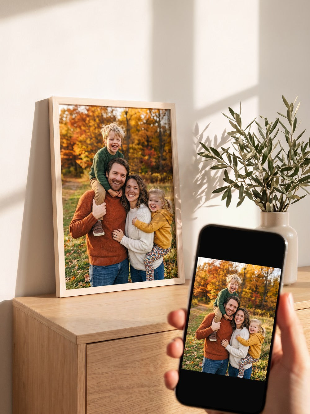 Familienfoto als Fotodruck im Holzrahmen Natur hell auf einem Sideboard, davor dasselbe Foto auf dem Handy