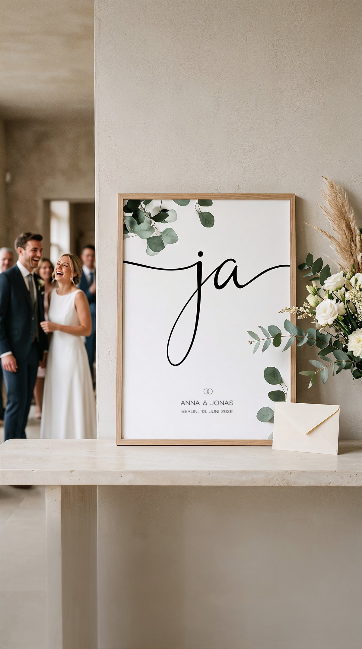 Gerahmtes Ja-Poster mit Eukalyptus auf dem Hochzeitstisch, dahinter unscharf ein lachendes Brautpaar