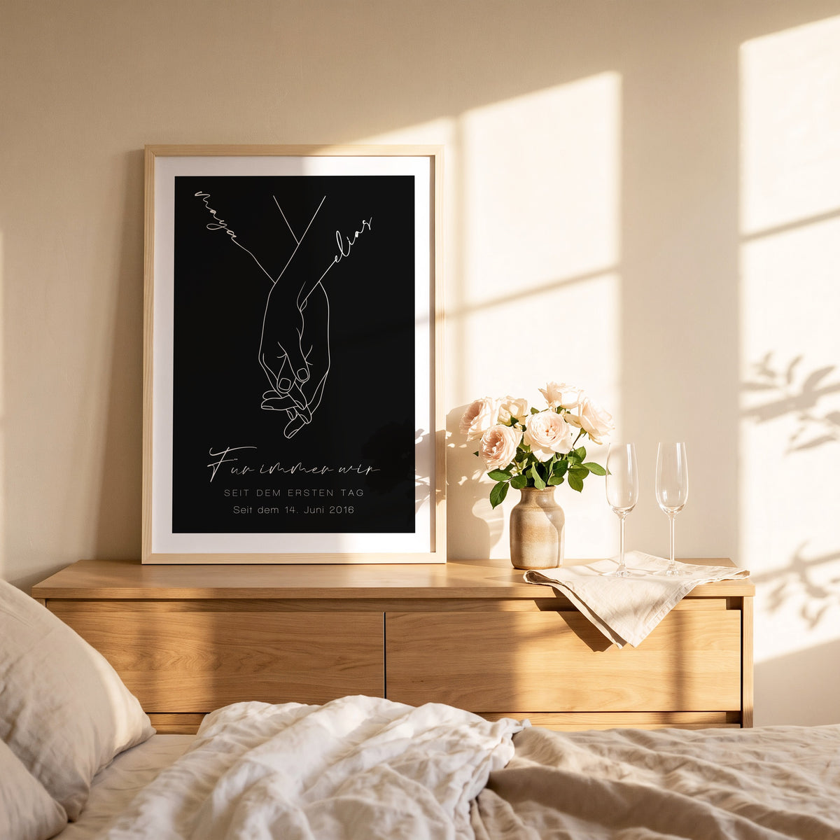 Schwarzes Hand-in-Hand-Poster mit Maya und Elias im Naturholzrahmen auf einer Kommode im Schlafzimmer
