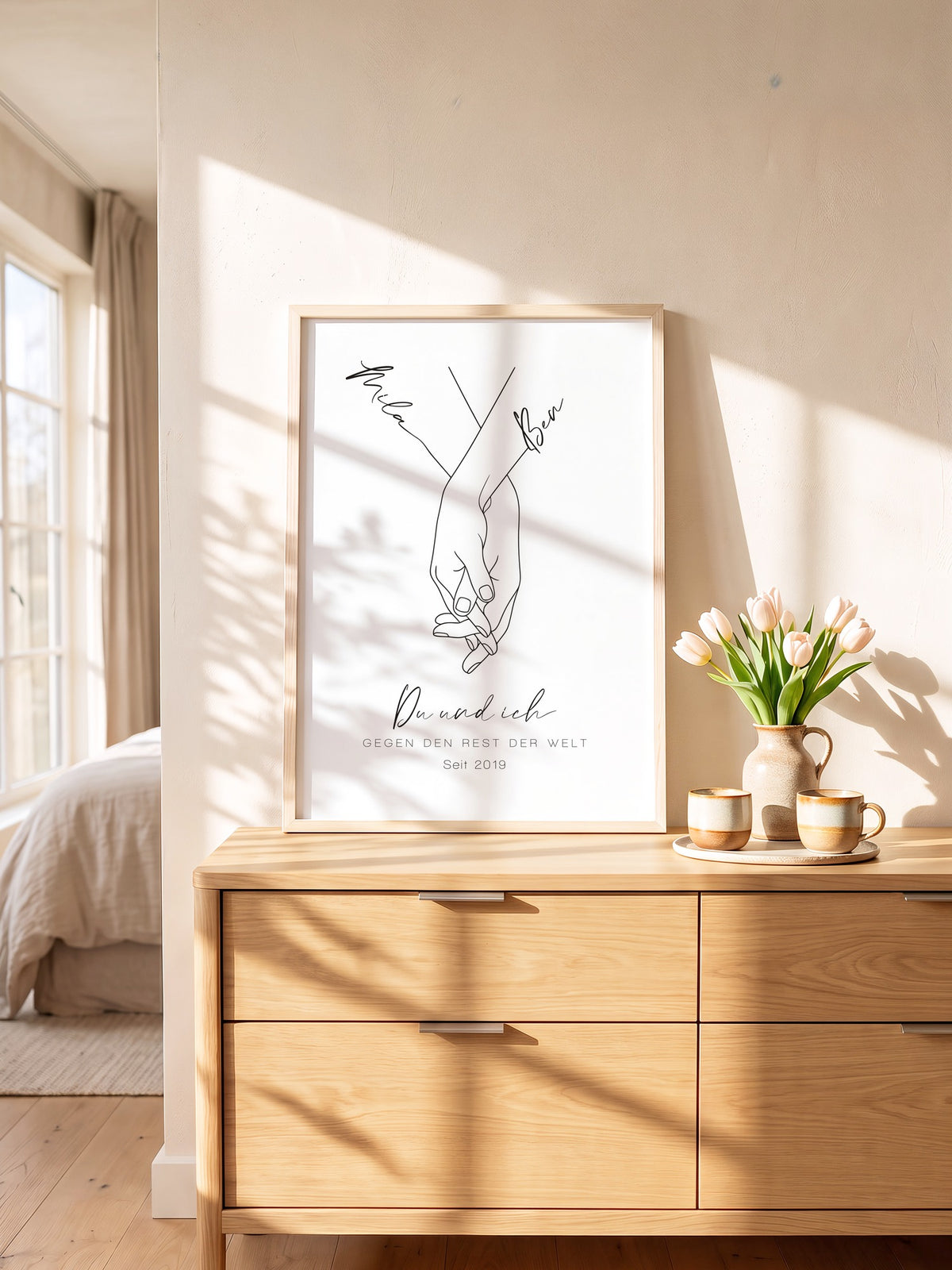 Weißes Hand-in-Hand-Poster mit Mila und Ben im Naturholzrahmen auf einer Kommode mit Tulpen