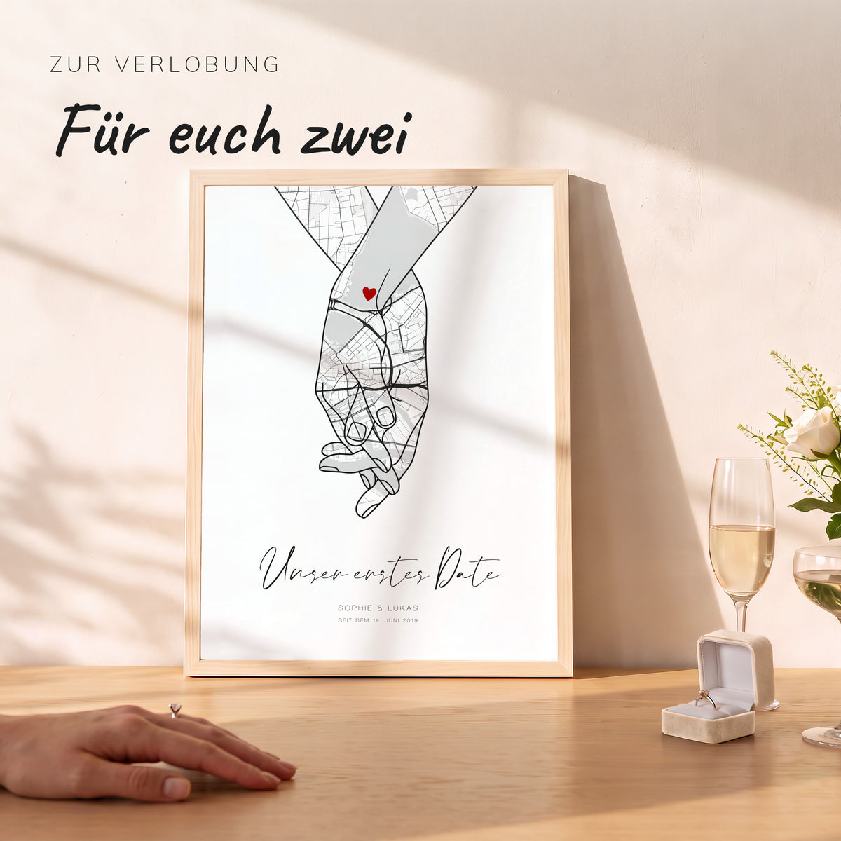 Hand-in-Hand-Kartenposter Unser erstes Date im Naturholzrahmen, davor Sektglas, Ringschachtel und eine Hand mit Verlobungsring