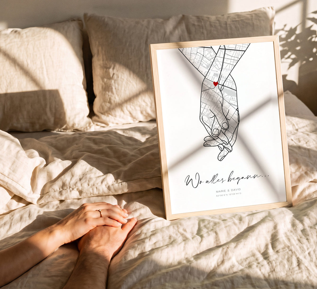 Hand-in-Hand-Kartenposter im Naturholzrahmen auf dem Bett, davor zwei Hände, die sich halten