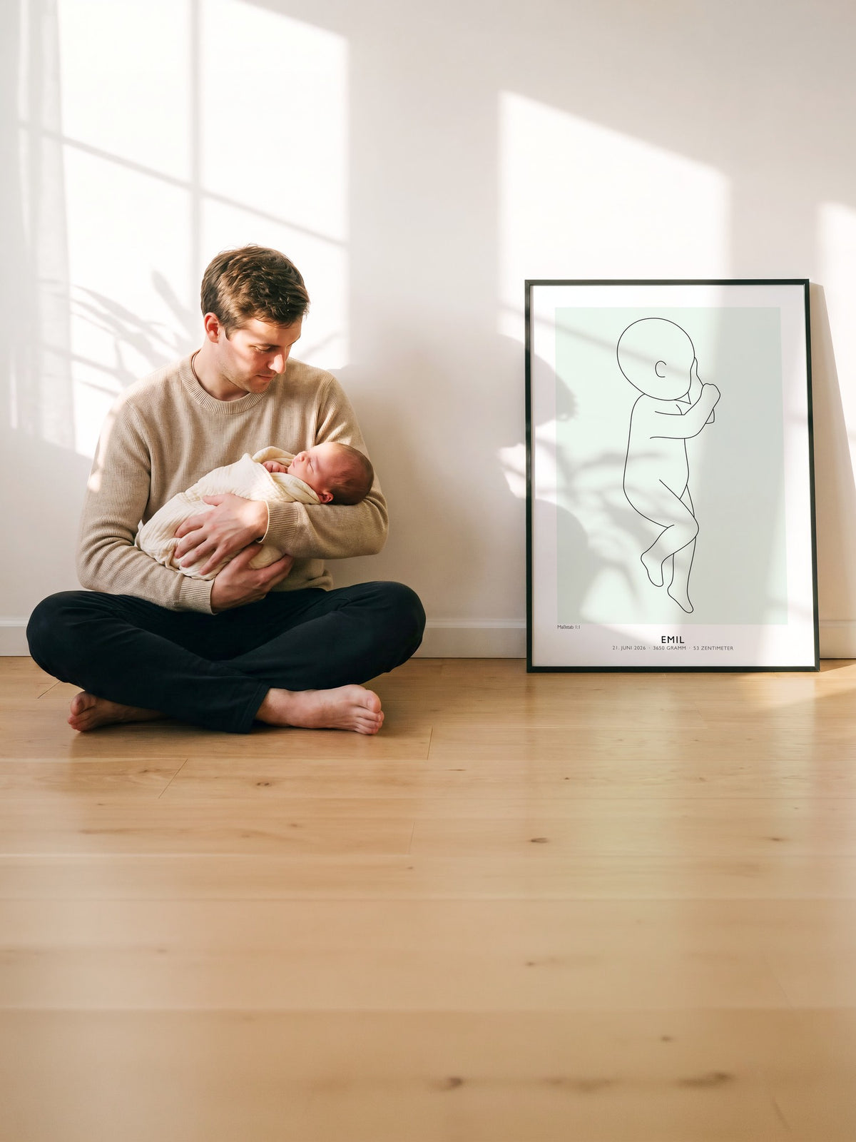 Vater sitzt mit seinem Neugeborenen im Arm an der Wand, daneben lehnt das gerahmte Geburtsposter von Emil in 50 × 70 cm
