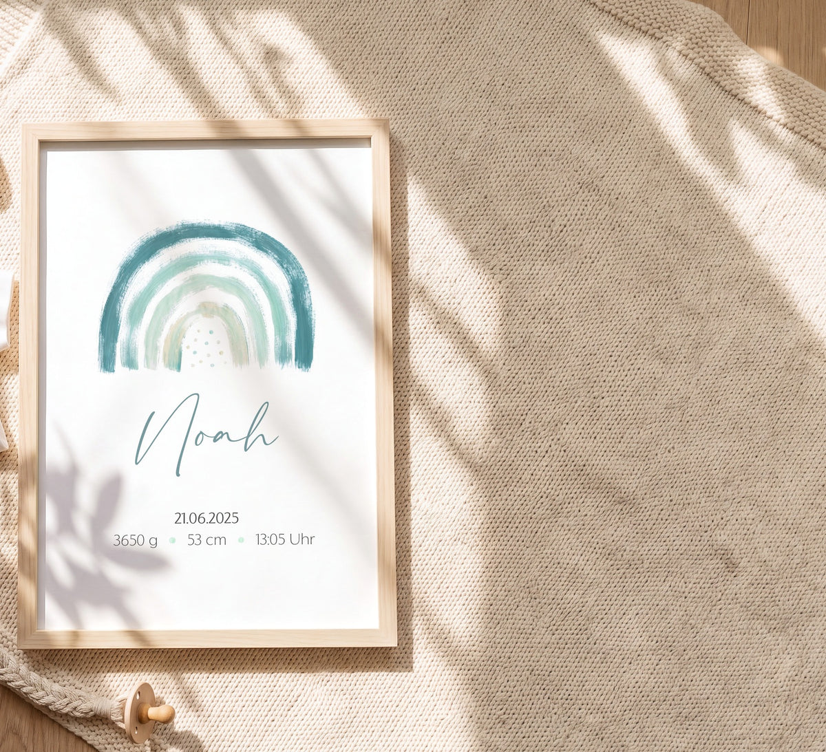 Geburtsposter mit grünem Regenbogen im hellen Holzrahmen auf einer Strickdecke neben Babybody, Mütze und Beißring