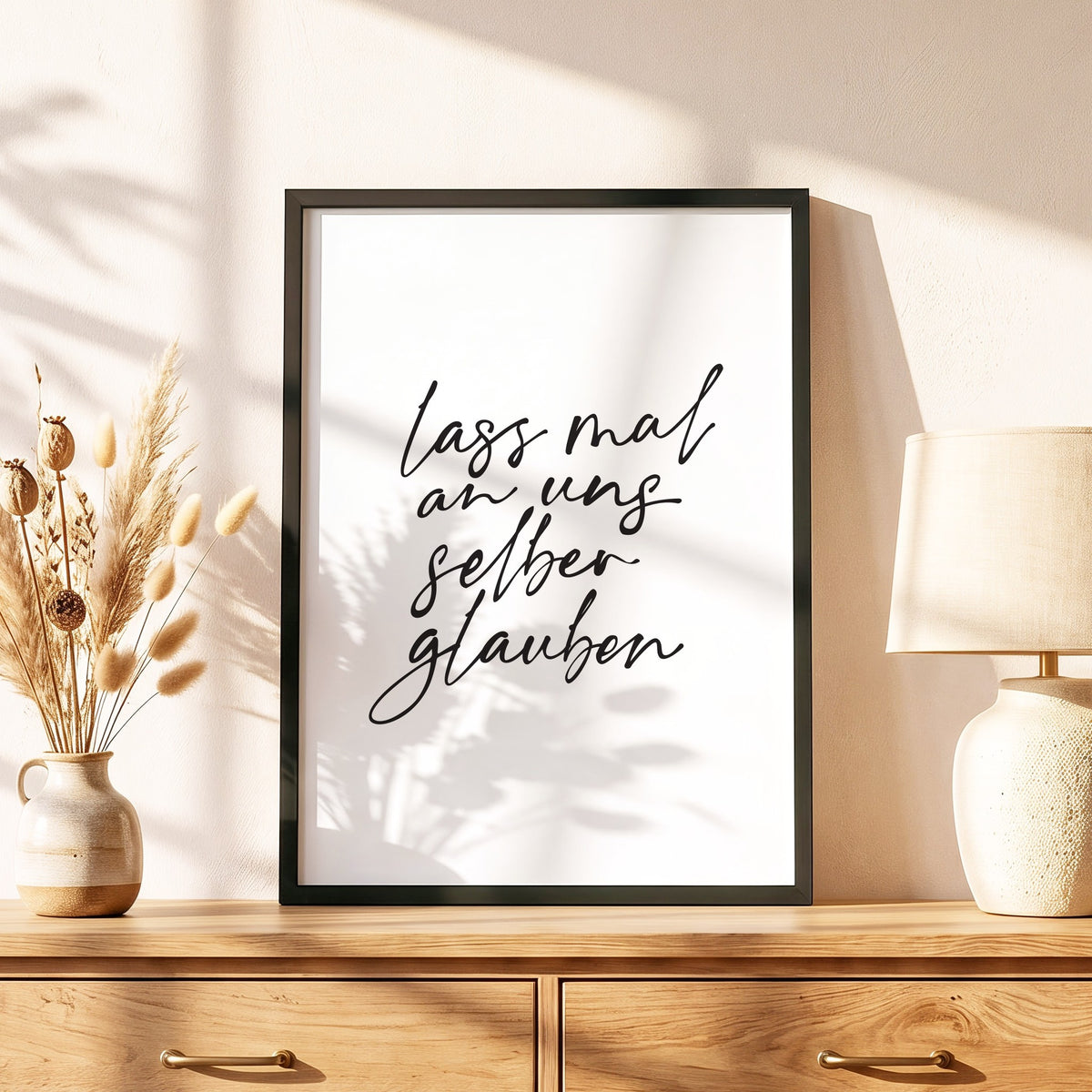 Spruchposter Lass mal an uns selber glauben im schwarzen Holzrahmen auf einer Kommode