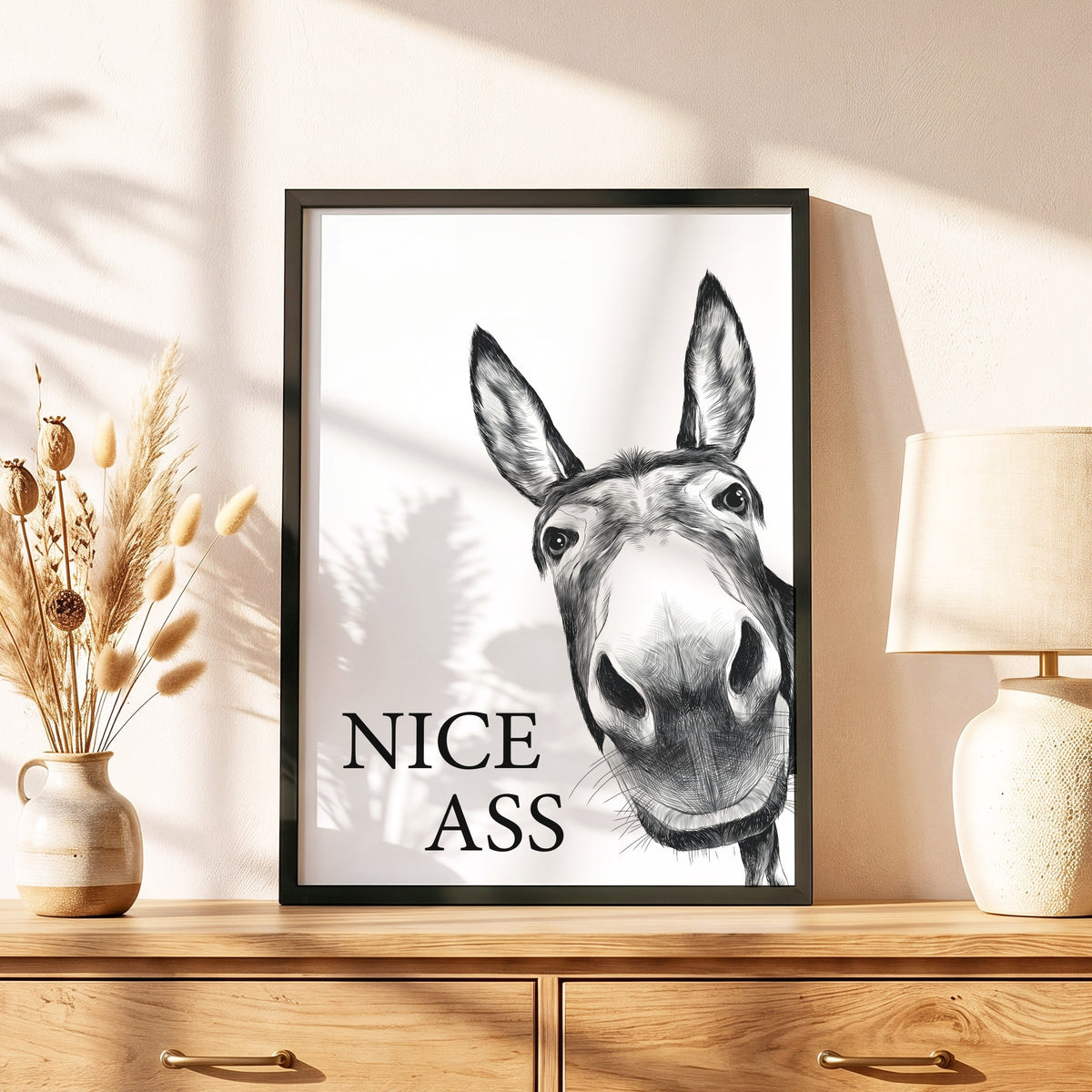 Spruchposter Nice Ass mit Esel im schwarzen Holzrahmen auf einer Kommode