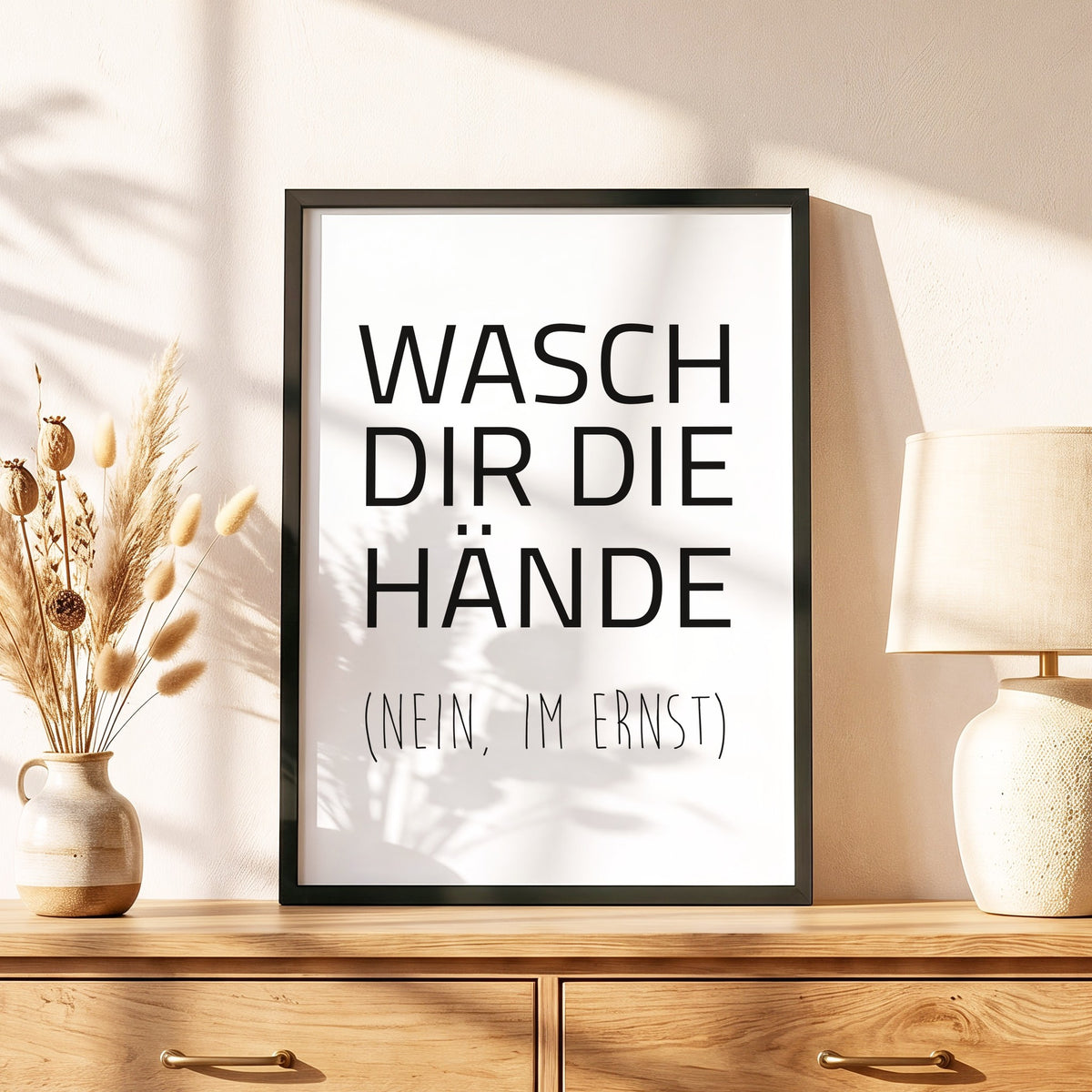 Spruchposter Wasch dir die Hände im schwarzen Holzrahmen auf einer Kommode