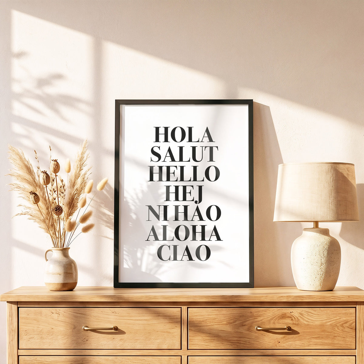 Poster Hola Salut Hello Ciao im schwarzen Holzrahmen auf einer Kommode